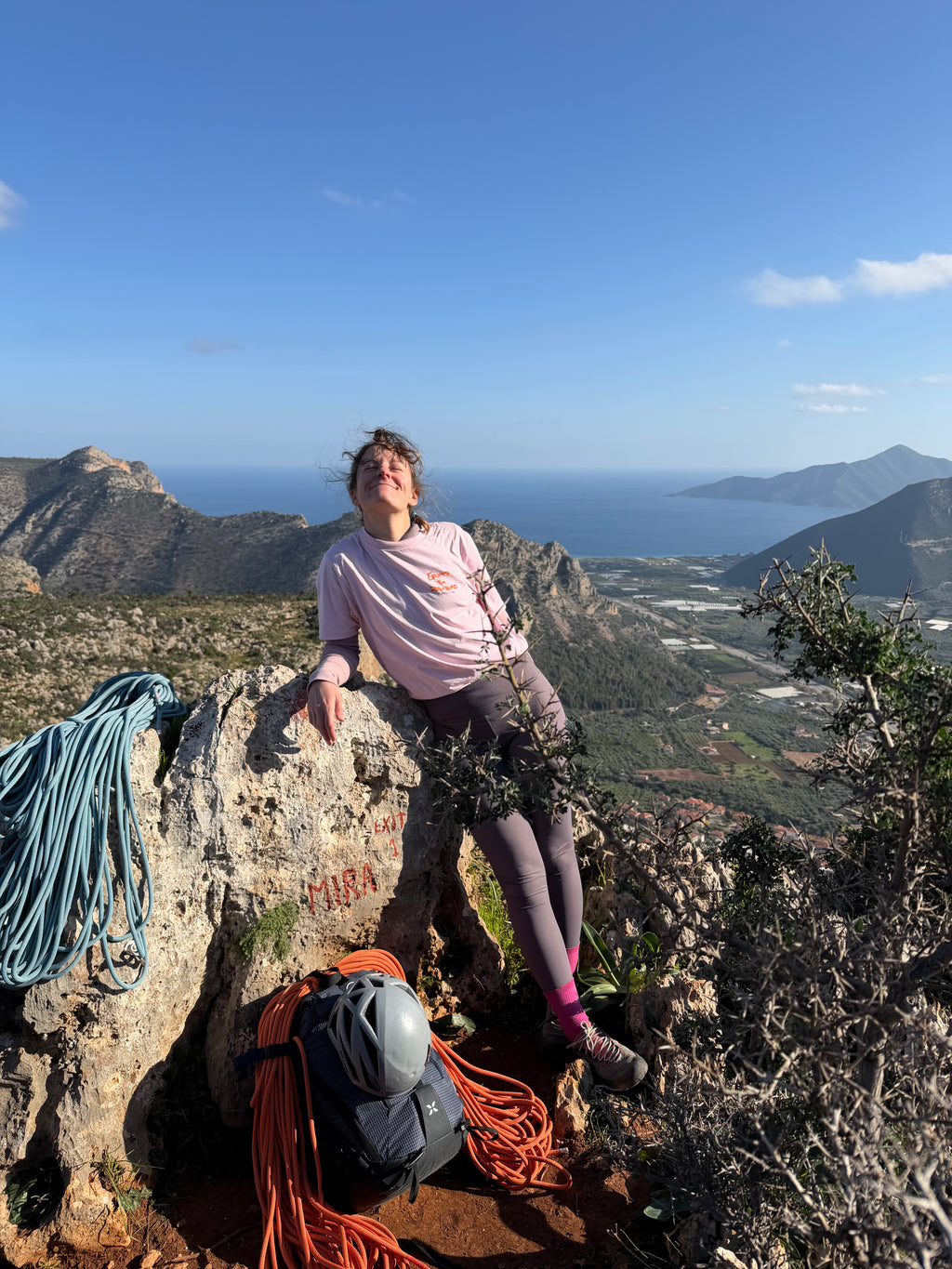 Person sitting on top of a multipitch in Greece with a scenic mountain and sea view in the background and climbing gear and the pink vertical vibes t-shirt unisex crimps for desayuno 